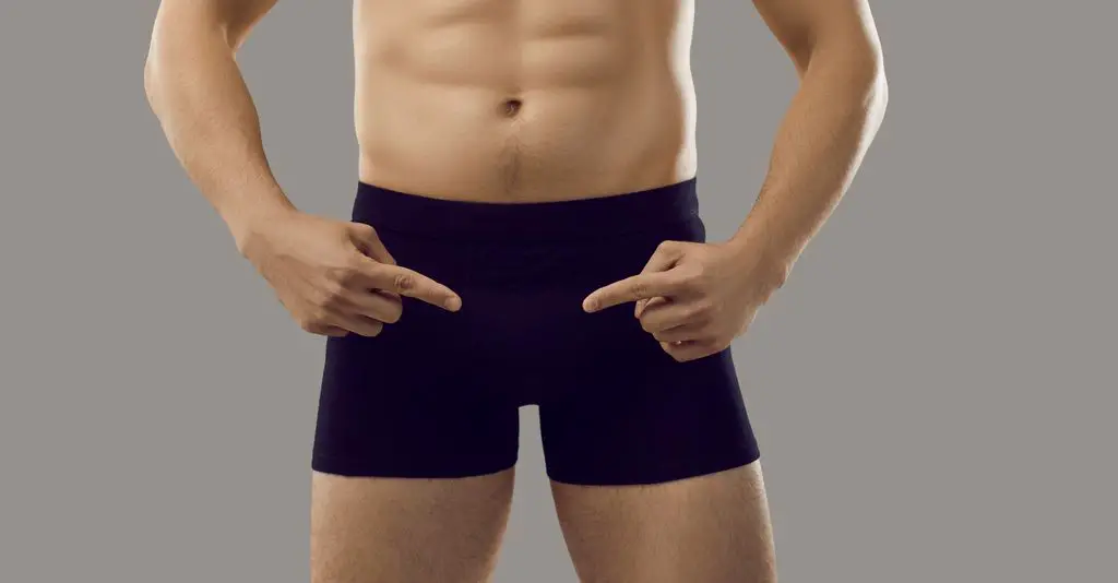Man fingers pointing at Contoured Pouch of black classic shorts boxers in which he is dressed, isolated on gray background.