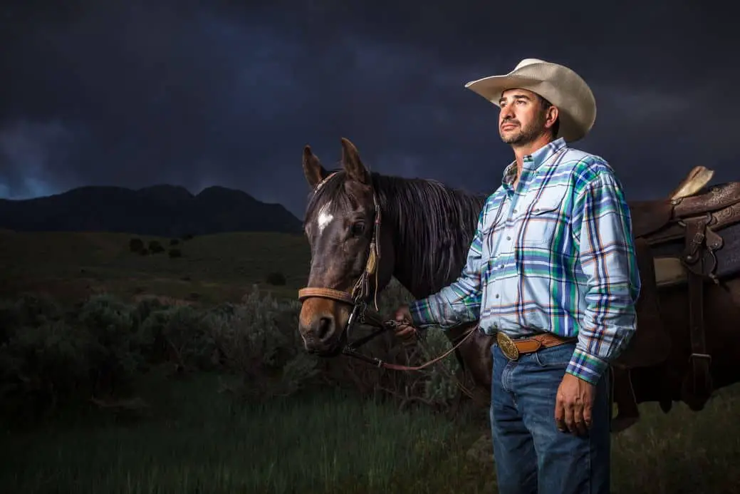 A man in his 30s wearing a checked shirt, blue jeans and a cowboy hat stands beside his horse on a rainy day., holding the reins. Dramatic portrait style shot taken outside in Goshen Canyon, Utah, 
