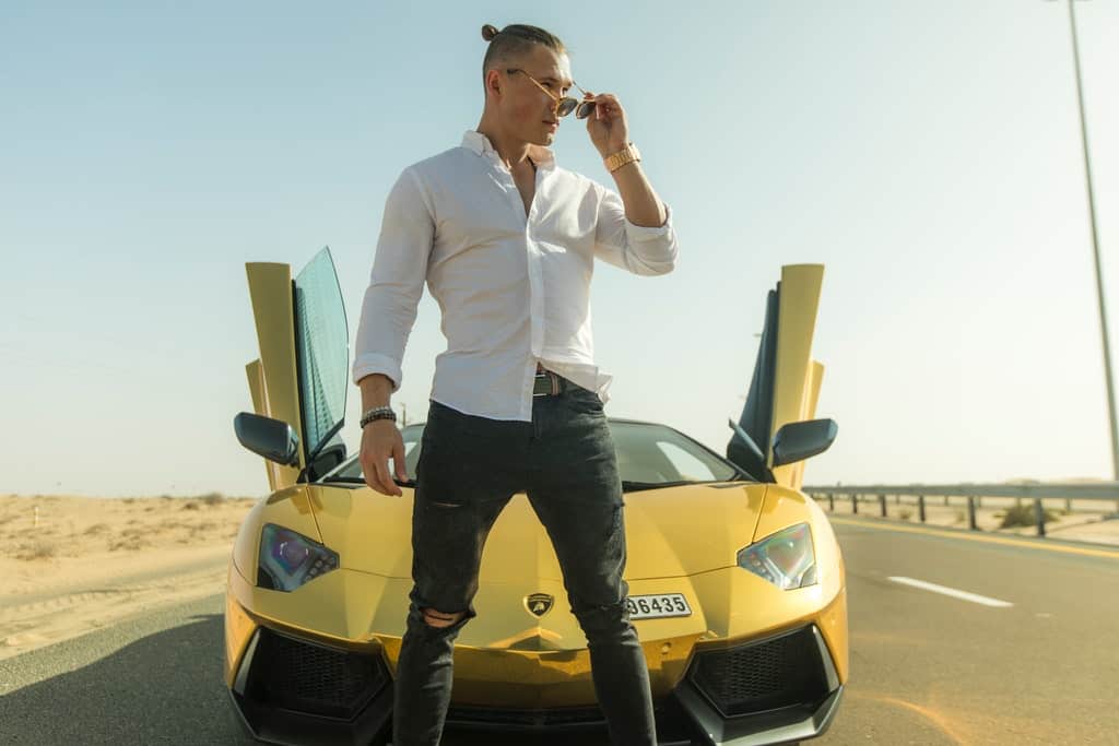 Serious young man in skinny jeans adjusting sunglasses and looking away near luxury car