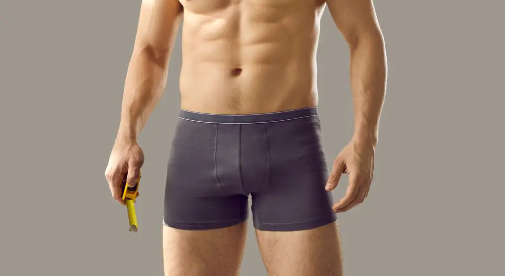 Does size matter. Man with fit, attractive body and naked torso measuring his big penis with centimeter tape. Man in boxer briefs holding yellow metal meter standing isolated on grey background